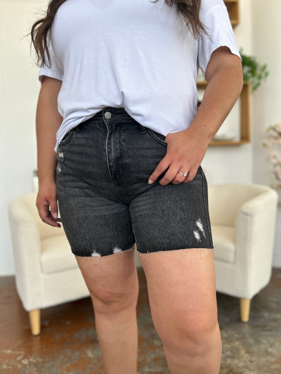 A person wearing a white t-shirt and Judy Blue Full Size High Waist Rigid Magic Denim Shorts is standing indoors in front of a beige chair.