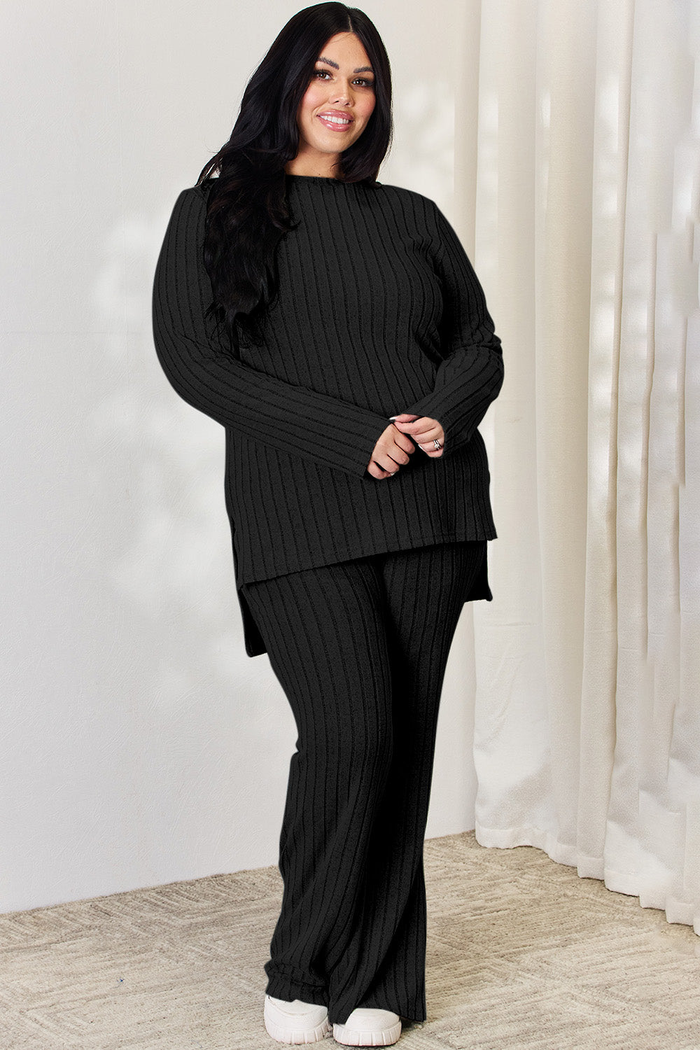 A person wearing the Basic Bae Full Size Ribbed High-Low Top and Wide Leg Pants Set in light gray, made from stretchy material, stands indoors in front of a white curtain.