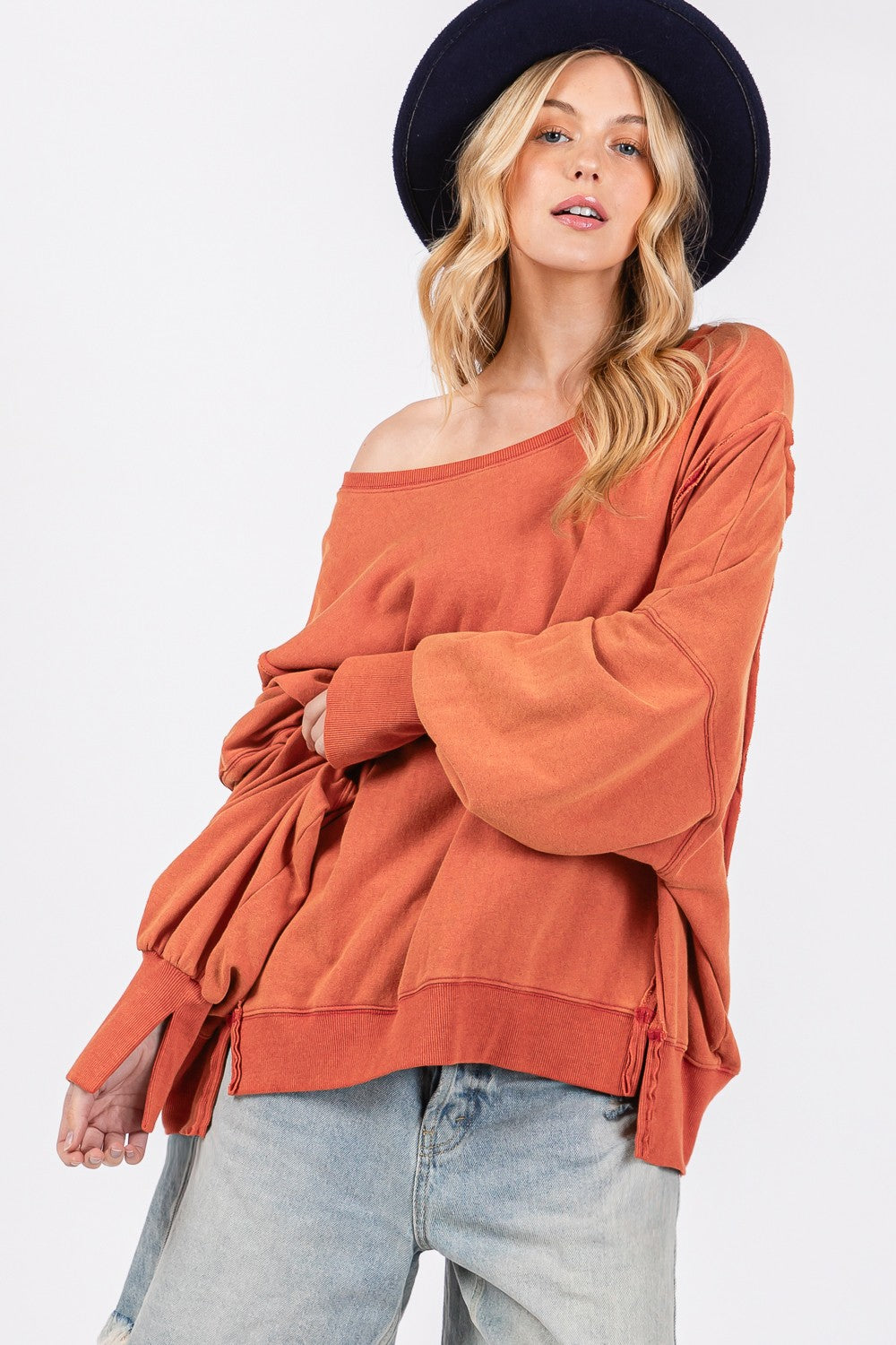 A person wearing a wide-brimmed hat, the SAGE + FIG Mineral Wash Side Slit Oversized Sweatshirt, and frayed denim shorts stands against a plain background.