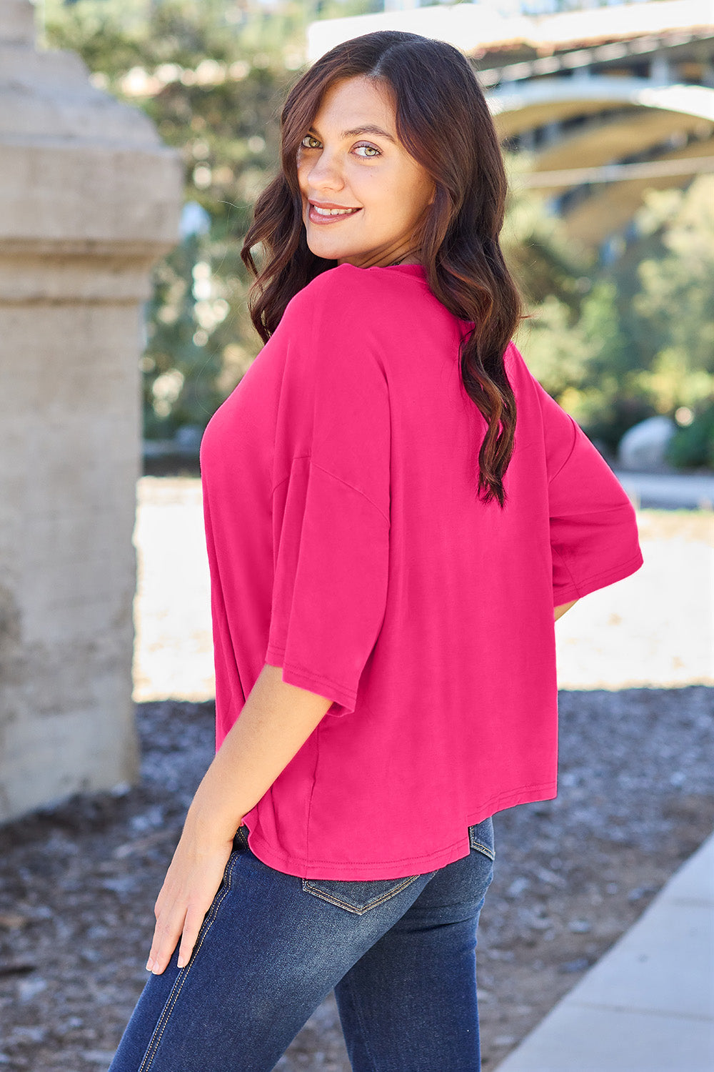 A person with long dark hair and a bright smile stands outdoors, showcasing their basic style in a loose, slightly stretchy *Basic Bae Full Size Round Neck Drop Shoulder T-Shirt* in pink and blue jeans, with one hand on their hip.