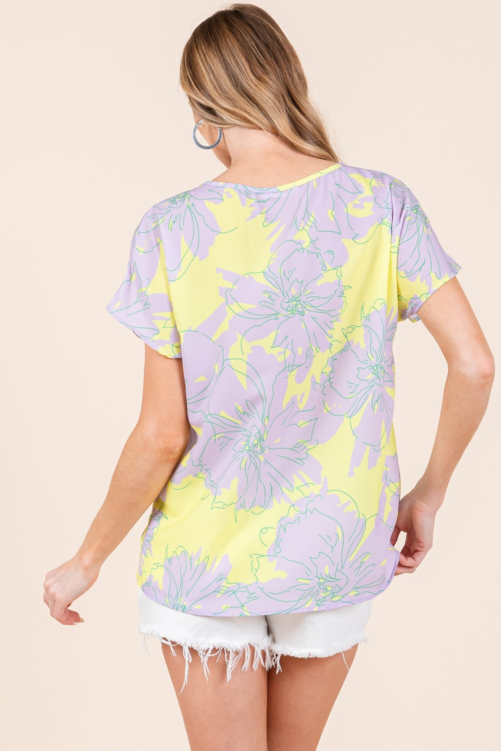 A woman wears the BOMBOM Floral Short Sleeve T-Shirt, featuring a vibrant floral print and a yellow V-neck, paired with white distressed shorts. She has long, wavy hair and stands against a beige background, showcasing her versatile and stylish look.