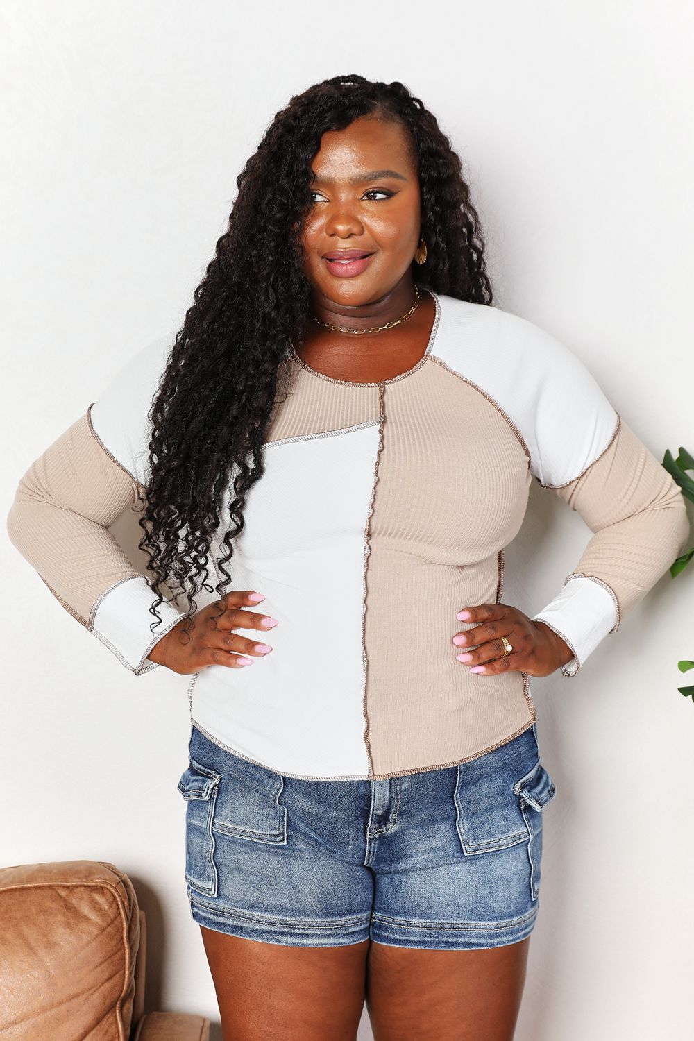 A woman with long brown hair is wearing the "Double Take Color Block Exposed Seam Top" in beige and white, paired with jeans. She stands with one hand in her pocket and the other touching her hair, smiling. A plant is visible in the background.