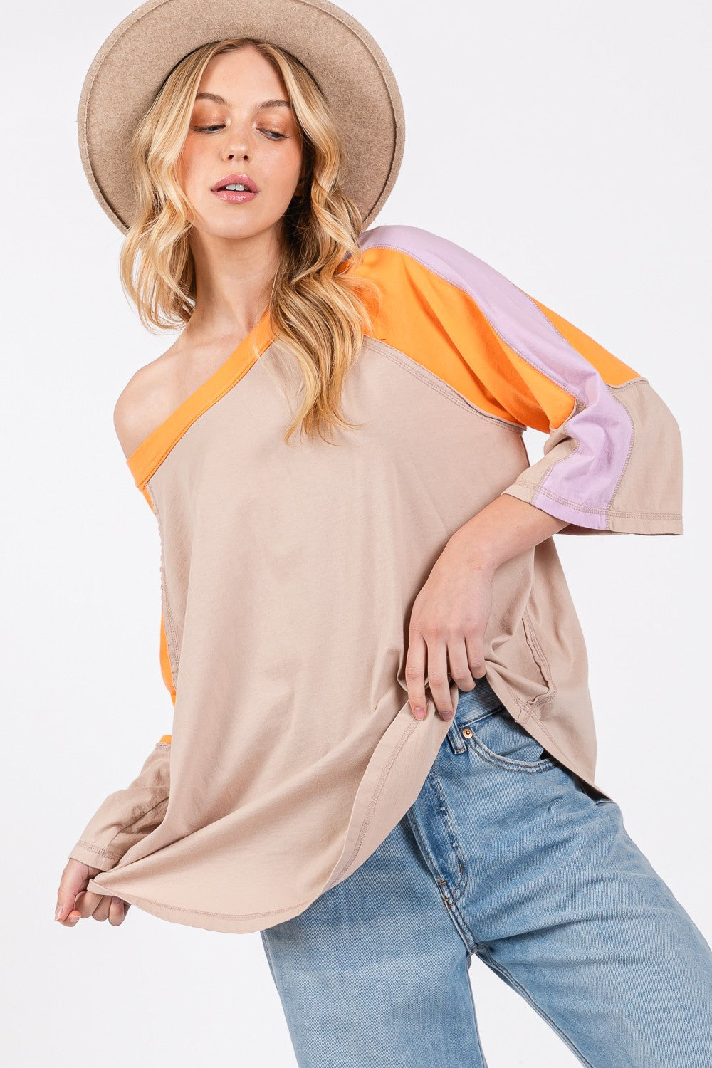 Person wearing a chic modern look with the SAGE + FIG Color Block Curved Hem T-Shirt in beige, featuring orange and purple accents, paired with blue jeans and a hat, poses against a plain background.