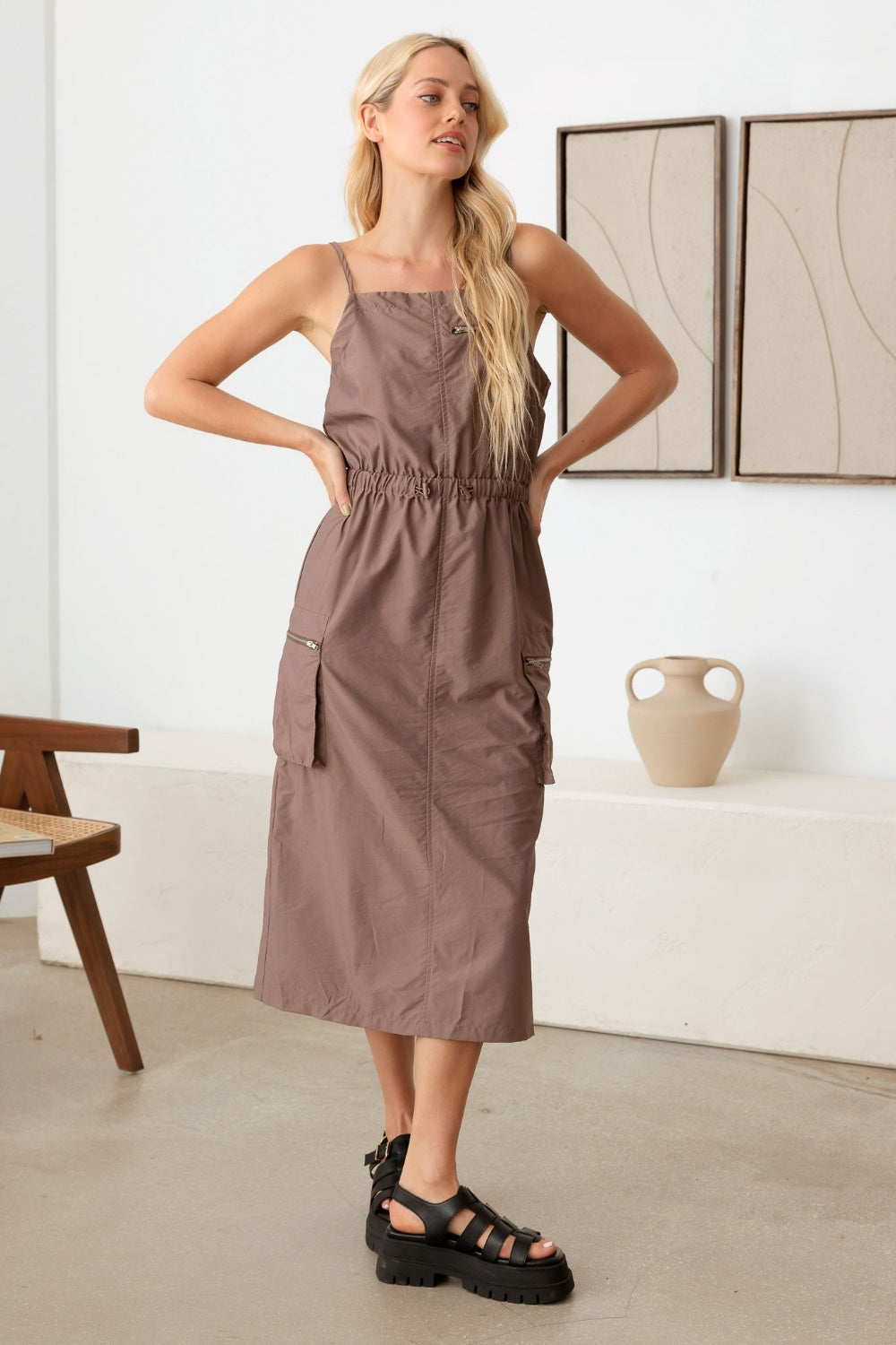 A woman stands indoors wearing the Le Lis Adjustable Strap Waist Drawstring Cargo Midi Dress paired with black platform sandals. She poses with her hands on her hips, showcasing the sleeveless brown dress with a cinched waist.