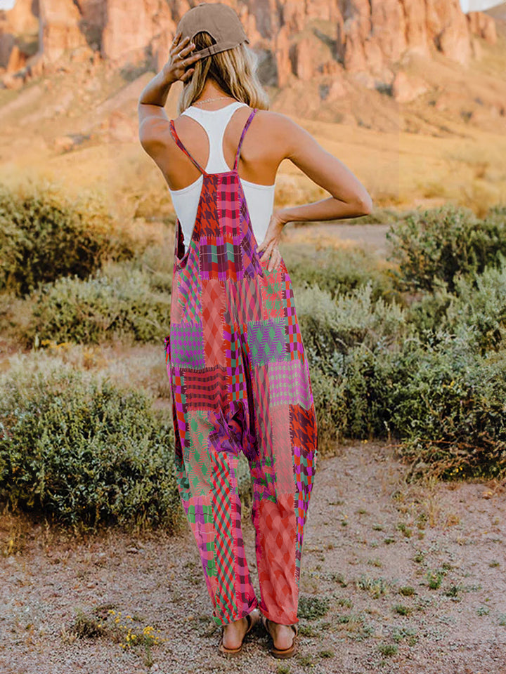 A woman stands outdoors in front of a rocky landscape, wearing the Full Size Printed V-Neck Sleeveless Jumpsuit and a brown brimmed hat. She is smiling and holding the brim of her hat with one hand, exuding the carefree vibe of this vibrant and colorful jumpsuit.