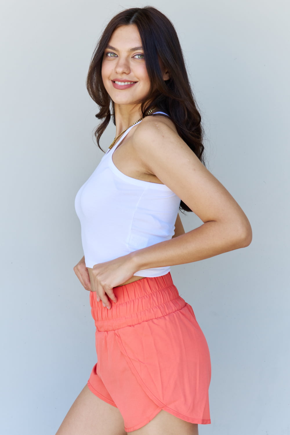 A person wearing the Ninexis Everyday Staple Soft Modal Short Strap Ribbed Tank Top in Off White, paired with coral shorts, stands smiling against a plain backdrop.