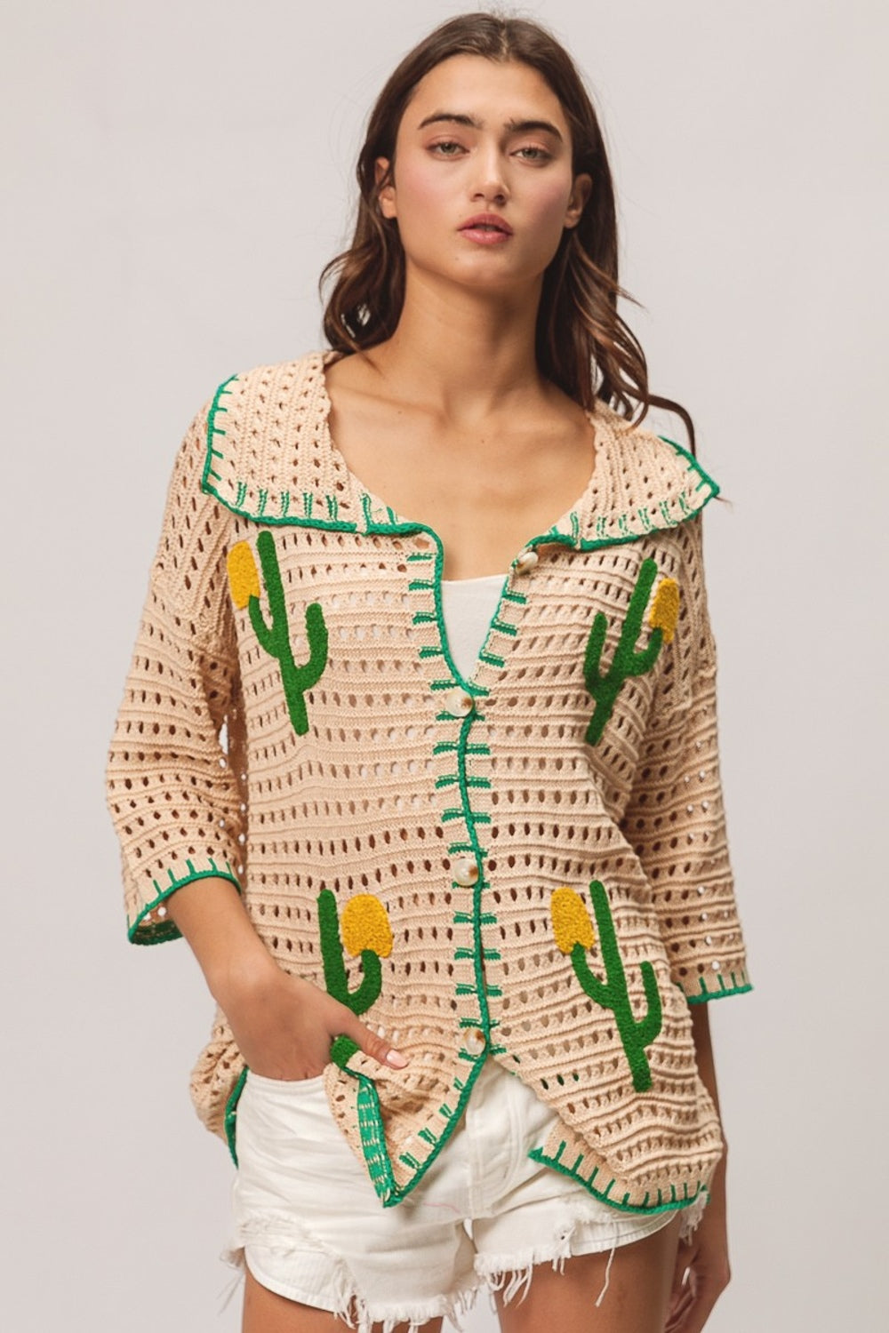 A woman wearing the BiBi Edge Stitched Cactus Patch Sweater Cardigan, featuring beige fabric with green cactus designs, layered over a white top and paired with white frayed shorts, stands against a plain background.