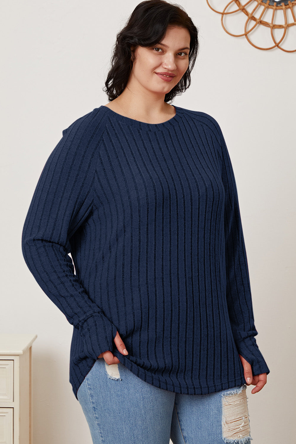 A person wearing the Basic Bae Full Size Ribbed Thumbhole Sleeve T-Shirt in blue and light-washed jeans stands smiling, with a decorative wall hanging in the background.