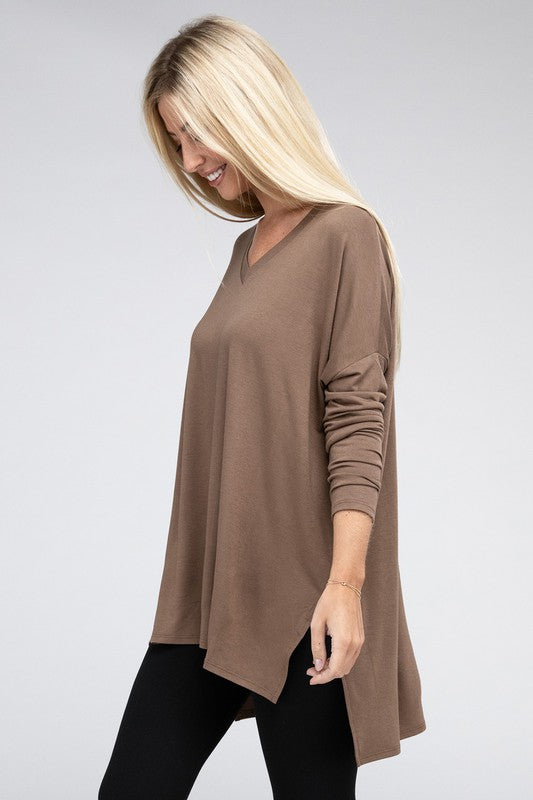 A woman with long, blonde hair is wearing a loose-fitting, brown Dolman Long Sleeve V-Neck Side Slit Hi-Low Hem Top and black pants. She poses with one hand on her hip and a neutral facial expression against a light grey background.