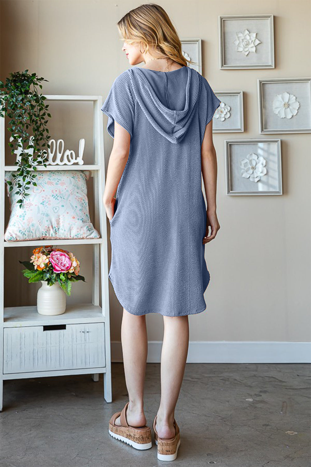 A woman wearing the Heimish Full Size Ribbed Short Sleeve Hooded Dress in a relaxed blue fit stands next to a window, touching her hair. A shelf with decorative items is visible in the background.