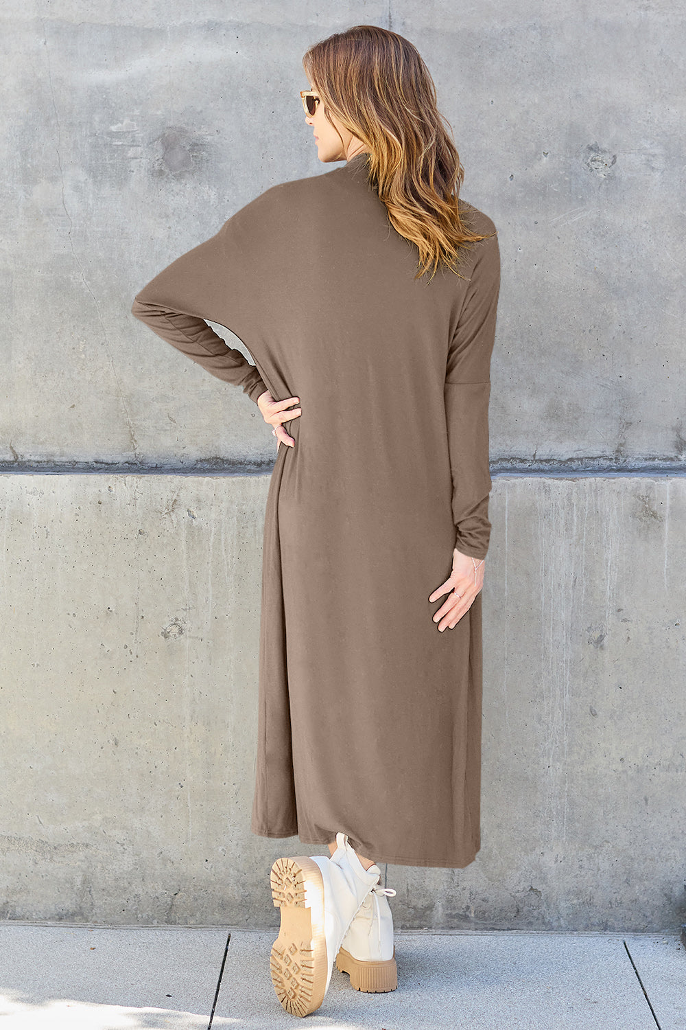 A woman stands against a concrete wall, wearing the Basic Bae Full Size Open Front Long Sleeve Cover Up made from rayon spandex, a white top, blue jeans in her perfect size, and brown shoes with her hands in her pockets. Machine wash cold for best results.