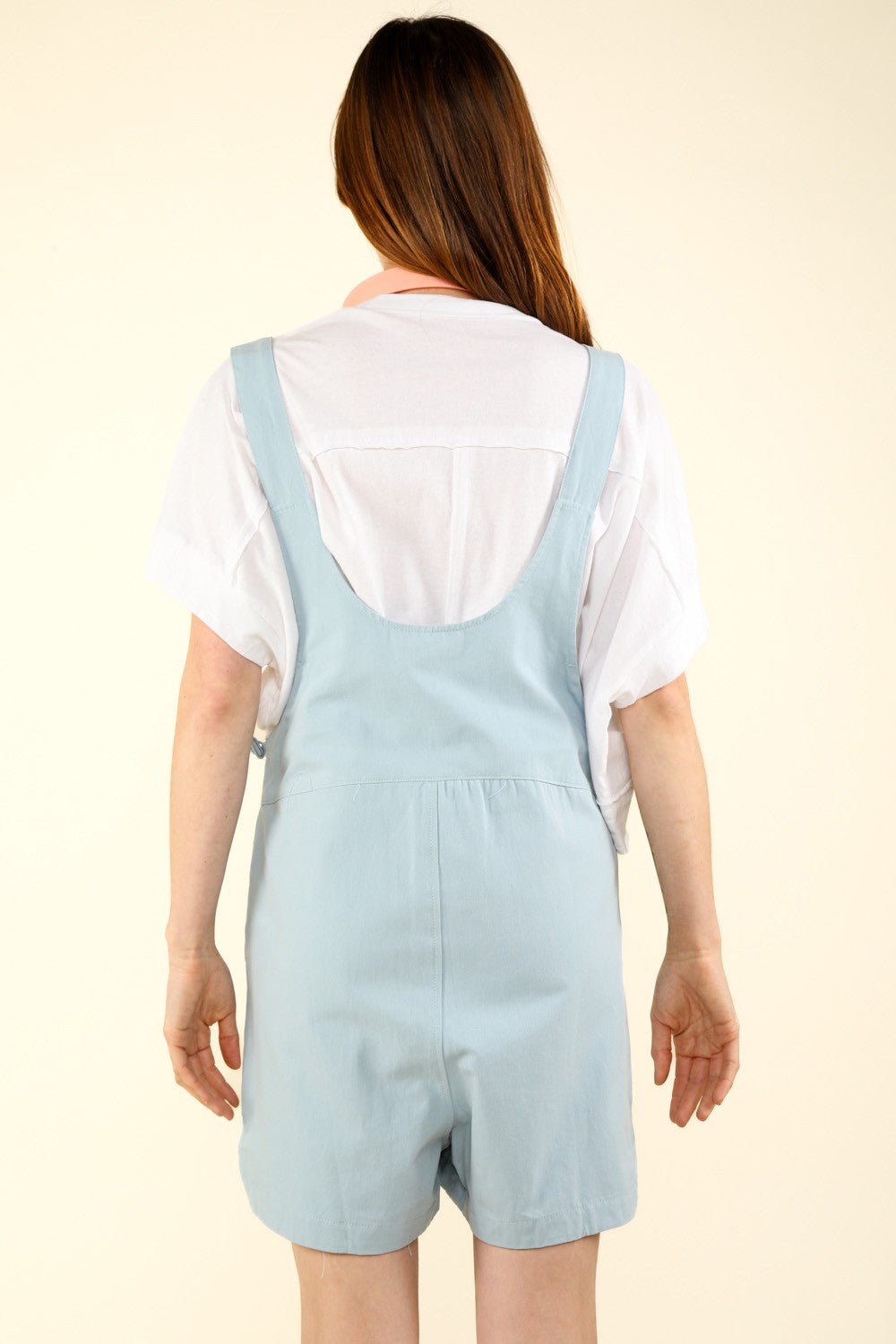 A person stands against a light background, wearing a white shirt, VERY J Adjustable Waist Suspender Overalls with Pockets, and pink headphones around their neck.