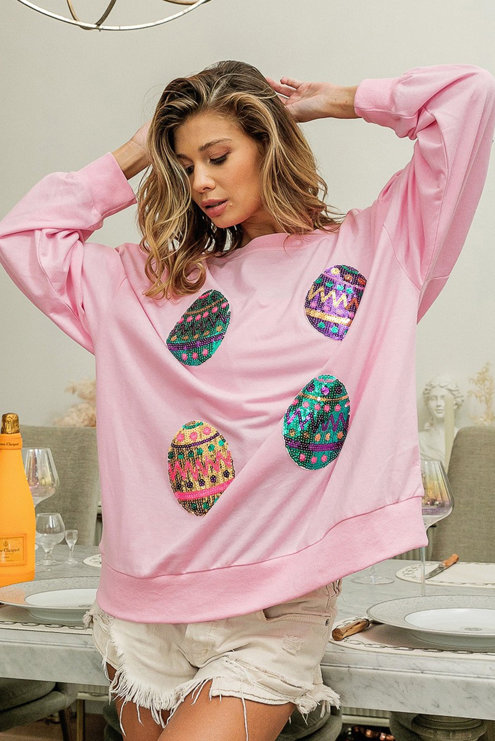 A woman with long, wavy hair wears a Pink Sequined Easter Egg Drop Shoulder Oversized Sweatshirt featuring colorful, patterned circles. She is seated in a dining area with a table set with glasses, plates, and two bottles, one of which is orange.