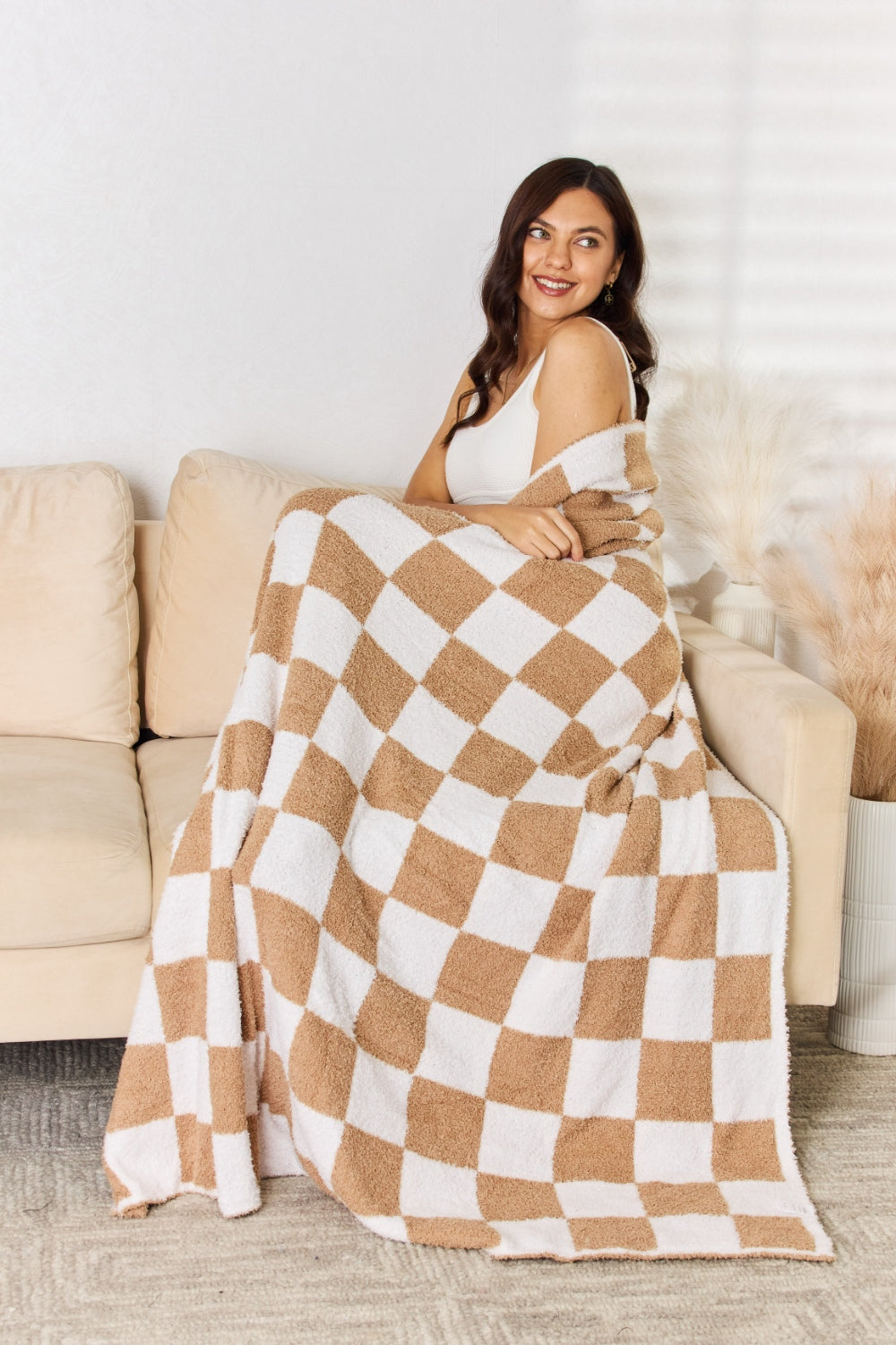 A person stands holding a large Cuddley Checkered Decorative Throw Blanket, smiling, in a room with a sofa and a potted plant. The blanket features a black and white checkerboard pattern, is made from 100% polyester, and imported for quality.