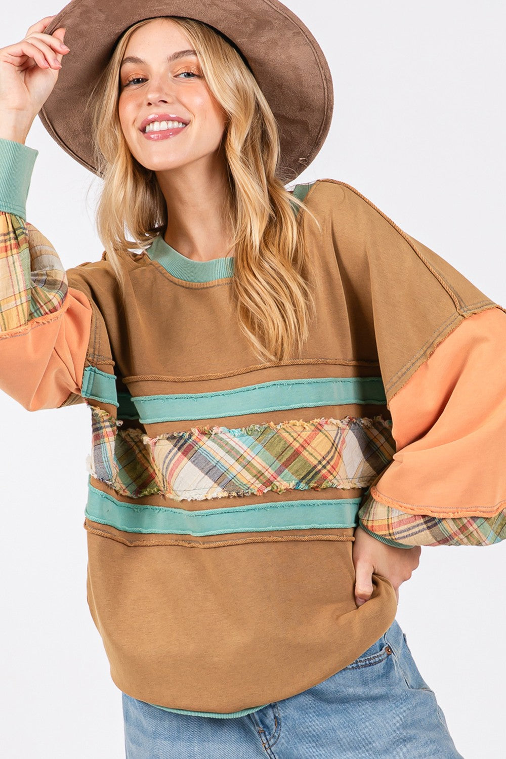 A person wearing a brown hat and the SAGE + FIG Mineral Wash Raw Edge Color Block Sweatshirt, featuring a patchwork design with plaid print contrast and solid color panels, smiles while posing with one hand in their pocket.