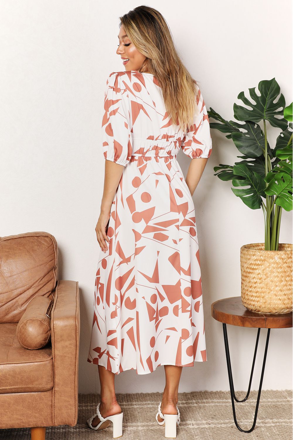 A person in a Printed Surplice Balloon Sleeve Dress stands near a brown chair and a round table with a potted plant.