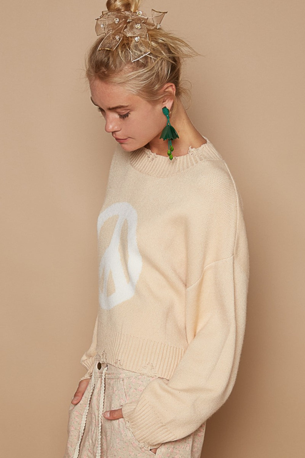 A woman smiles, donning the POL Round Neck Long Sleeve Peace Sign Sweater in beige, green tassel earrings, and beige patterned pants.