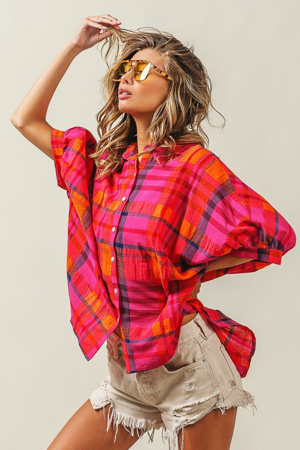 A woman outfitted in a BiBi Button Up Dolman Sleeve Plaid Shirt, paired with a white crop top, distressed denim shorts, and yellow sunglasses.