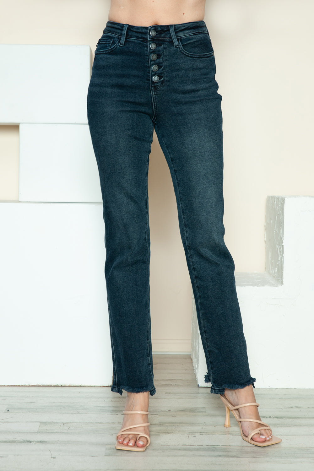 A person is modeling the Judy Blue Full Size Button Fly Hem Destroy Straight Jeans, high-waisted with distressed detailing and frayed hems, paired with light-colored open-toe heels. The background includes white blocks and a light-colored wall.