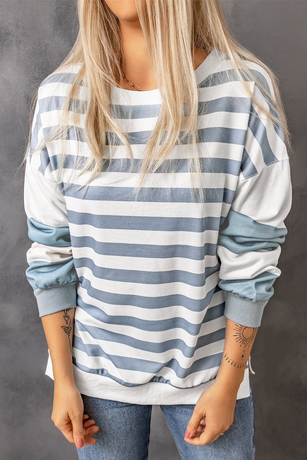 A person with long blonde hair, wearing a Stripe Drop Shoulder Striped Pullover Sweatshirt and relaxed-fit jeans, is standing facing a gray wall with their right hand raised. Their back is to the camera.