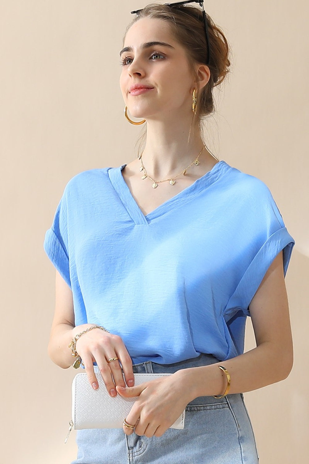 A woman wearing a stylish Ninexis V-Neck Trim Rolled Short Sleeve Shirt in light blue and high-waisted blue jeans, accessorized with hoop earrings, a necklace, and sunglasses on her head, poses in front of a beige background.