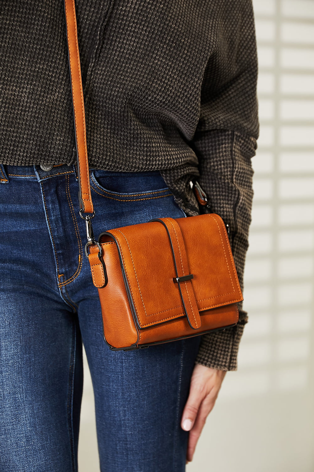 A person wearing a dark sweater and jeans holds the SHOMICO PU Leather Crossbody Bag, a stylish accessory in orange-brown featuring a flap and buckle detail.