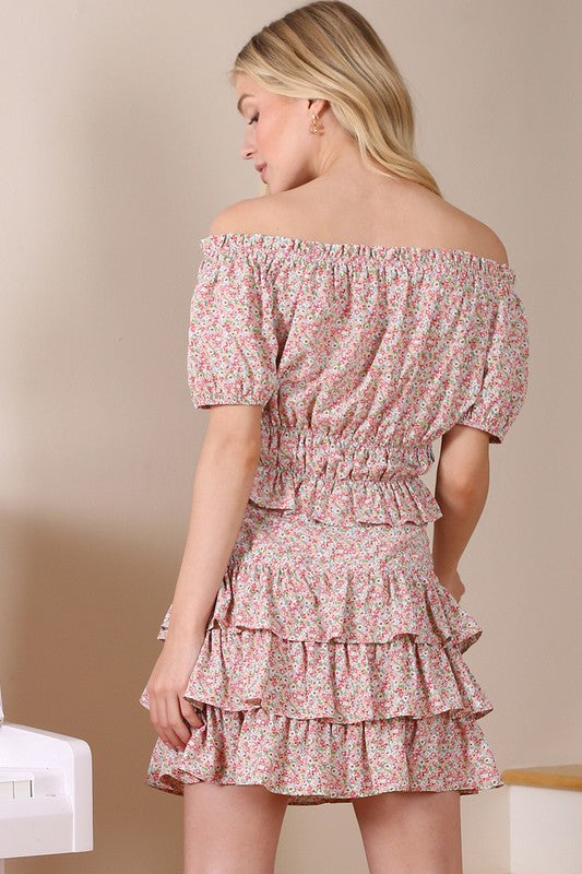 A woman wears the SL off shoulder tube top, featuring a floral print and a shirred design. The ensemble is completed with puffed sleeves and a ruffled skirt for an elegant and charming look.