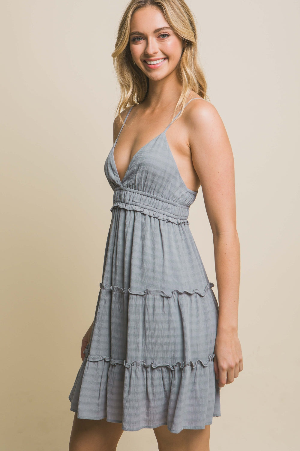 A woman in a light blue Love Tree Frill V-Neck Cami Dress with a tiered skirt stands against a beige background, holding one hand near her head and looking at the camera. This lightweight and flowy fabric is perfect for special occasions.