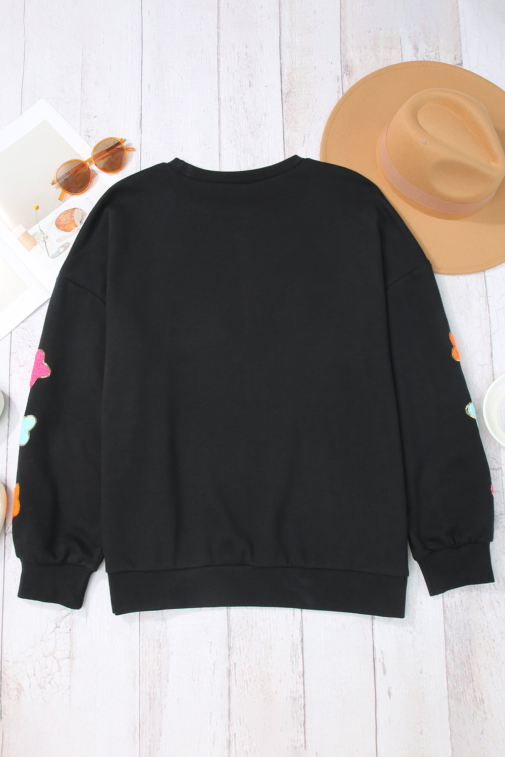 A woman with short blonde hair is wearing the Black Howdy Glitter Chenille Patch Graphic Casual Sweatshirt, featuring colorful star patterns and "WD" letters. She pairs it with light denim shorts and smiles while looking to the side.