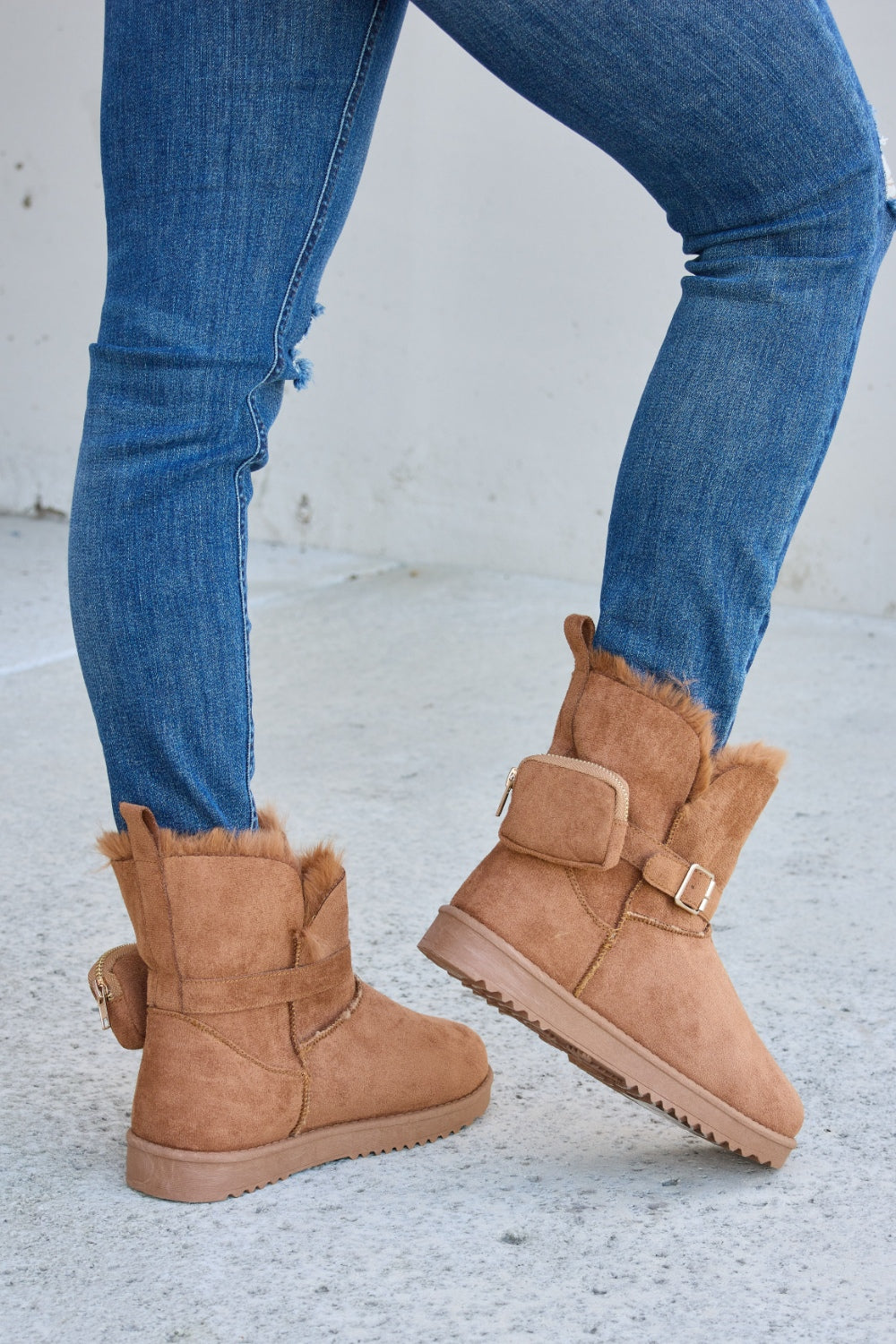 Person wearing ripped blue jeans and Forever Link Plush Thermal Flat Boots, tan in color, fur-lined with side buckles, standing on a concrete surface. These stylish boots promise cozy insulation for the feet.