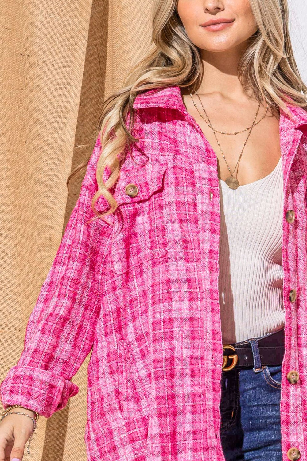 A woman stands smiling, wearing the "And The Why Full Size Tweed Button Down Long Sleeve Shacket" in pink plaid over a white top with a black belt and jeans. She is against a wall partially covered by a burlap curtain.