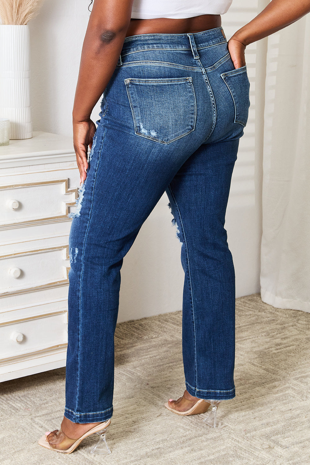 A person wearing Judy Blue Full Size Mid Rise Hand Sand & Destroy Bootcut Jeans stands with hands in pockets. The slightly stretchy jeans have rips on both legs, and the person is standing indoors beside a white dresser.