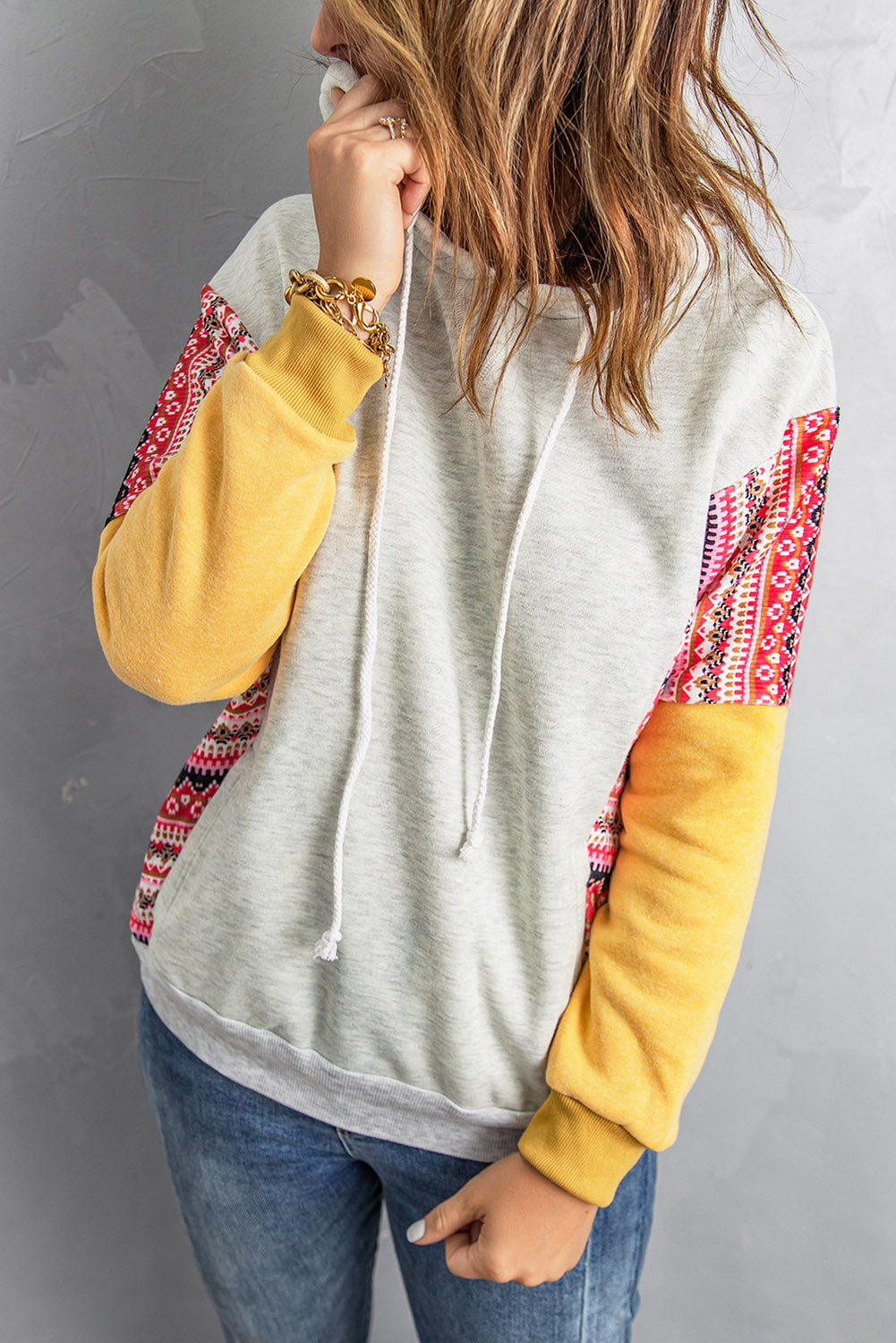 A person with long hair wearing an Aztec Patch Pullover Hoodie and blue jeans is facing away from the camera against a textured gray wall.