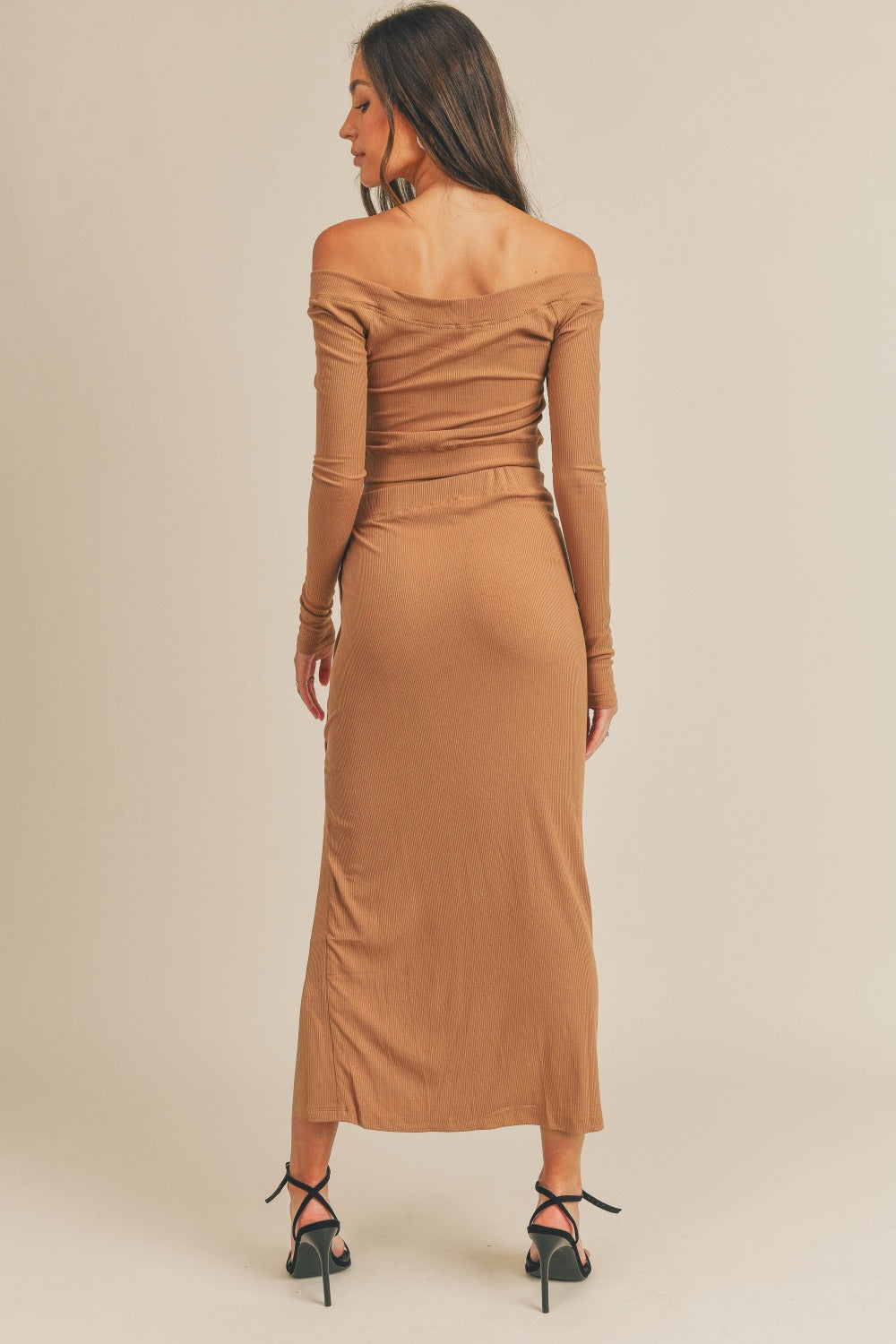 A woman stands against a plain background wearing the MABLE Button Detail Crop Top and Slit Midi Skirt Set, showcasing a camel-colored ensemble with off-shoulder, button details, and a side slit. She completes her look with black high-heeled sandals.