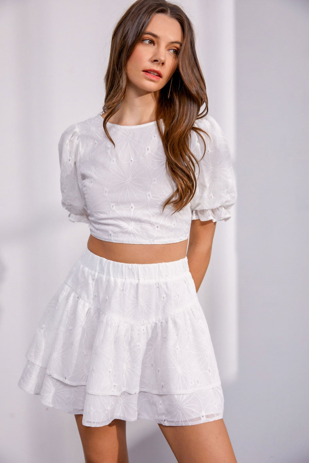 A woman wearing the Mustard Seed Floral Embroidered Back Lace Up Crop Top, which is adorned with floral embroidery and paired with a matching tiered skirt featuring subtle patterns, stands against a light background.