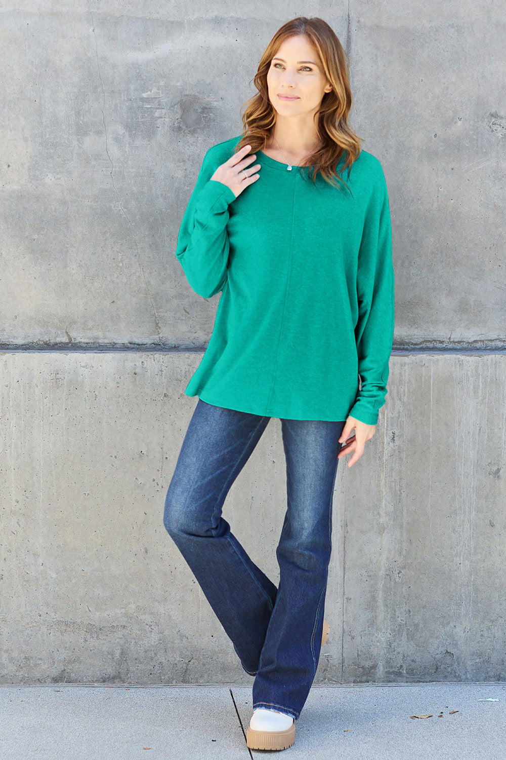 A person wearing a Double Take Full Size Round Neck Long Sleeve T-Shirt in green, blue distressed jeans with a slightly stretchy fit, and a crossbody bag, holding a beverage in a plastic cup.