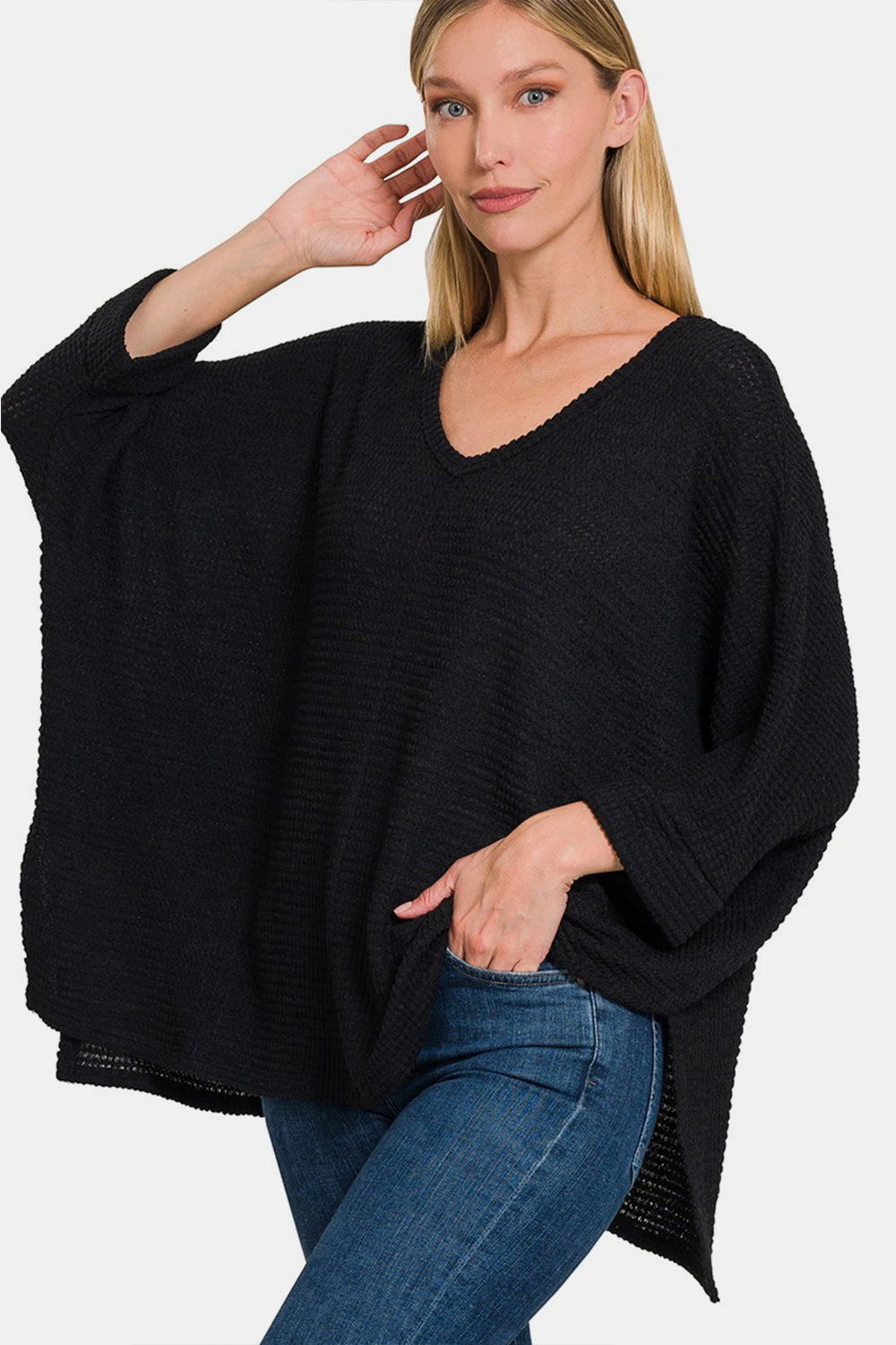 A woman wearing the Zenana Full Size Waffle Knit V-Neck Long Sleeve Slit Top paired with blue jeans stands with one hand touching her head and the other in her pocket.