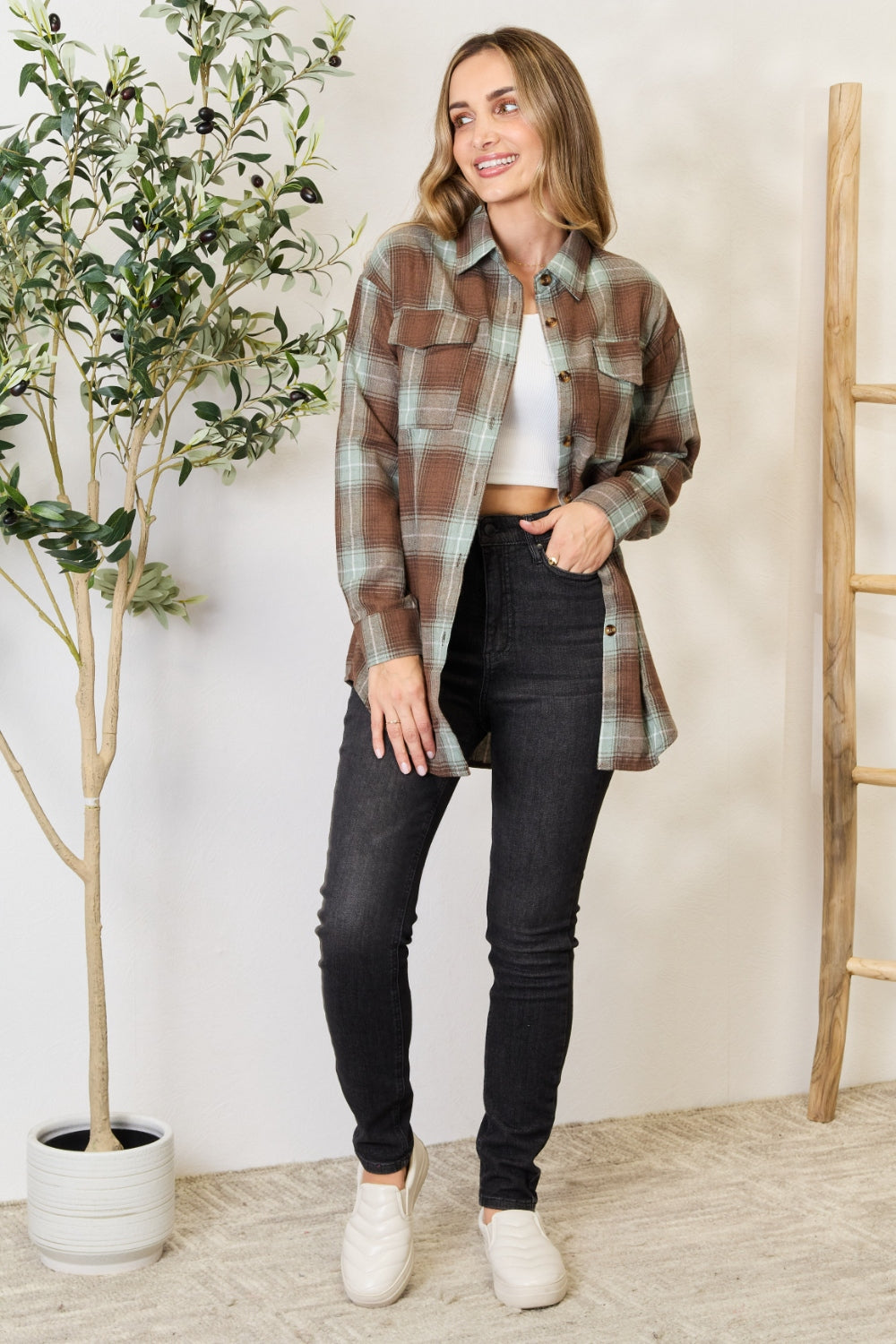 A woman stands in front of a light-colored wall and an olive tree, wearing the Mandy Plaid Dropped Shoulder Shirt paired with a white crop top and dark jeans. She smiles and holds her hair back with one hand.