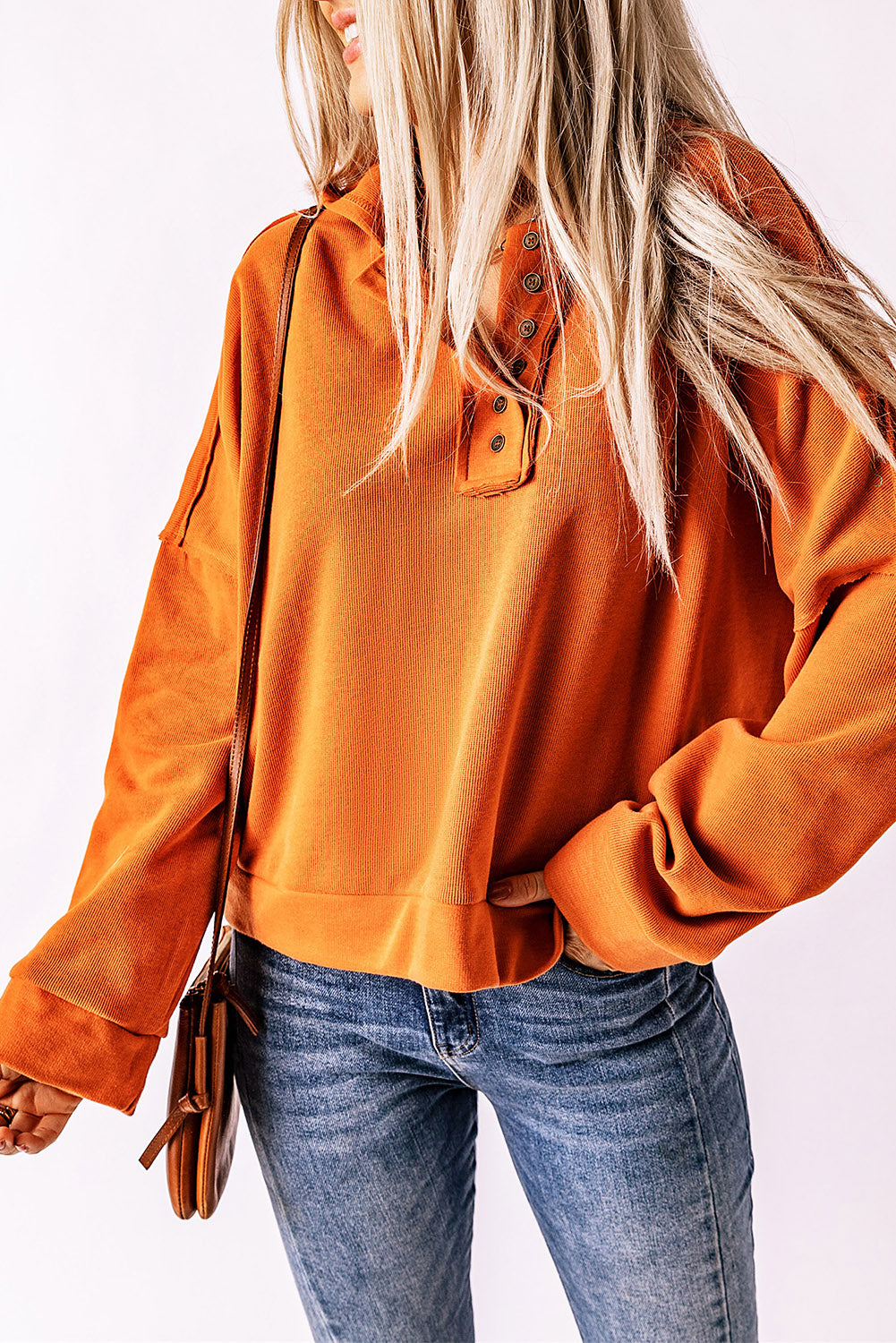 A person with long blonde hair, dressed in the Orange Casual Button Solid Patchwork Trim Hoodie and blue jeans, is holding the hood of their hoodie with one hand while facing away from the camera. This outfit is ideal for fall and winter essentials.