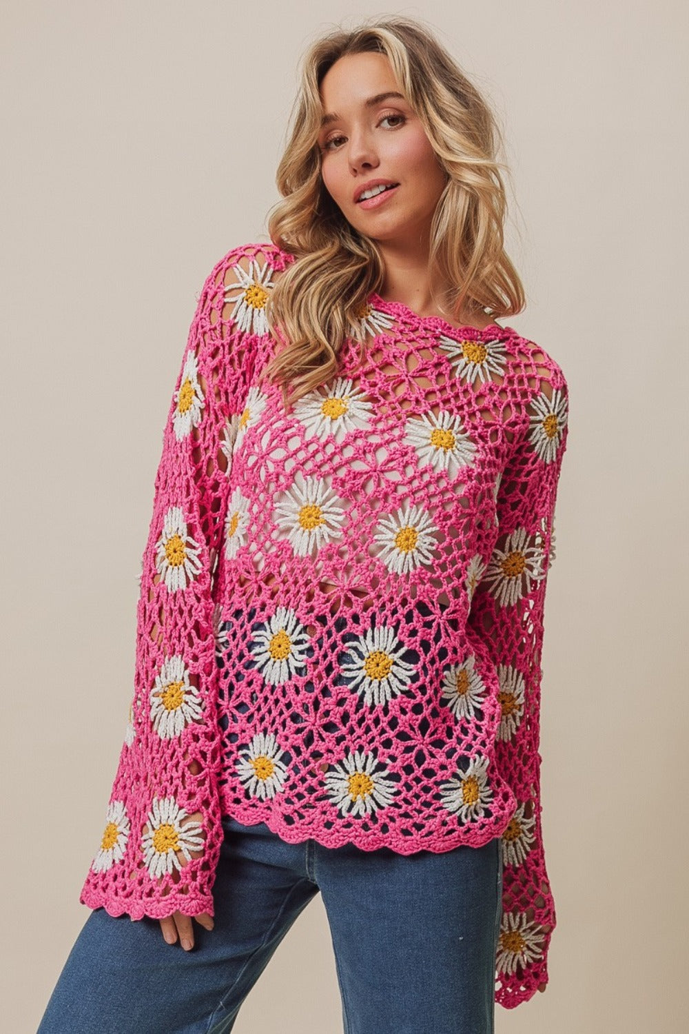 A person stands wearing the BiBi Floral Crochet Net Lace Cover Up, featuring pink crochet work adorned with white and yellow daisy patterns, exuding Bohemian style. They have long, wavy hair and pose with hands together.