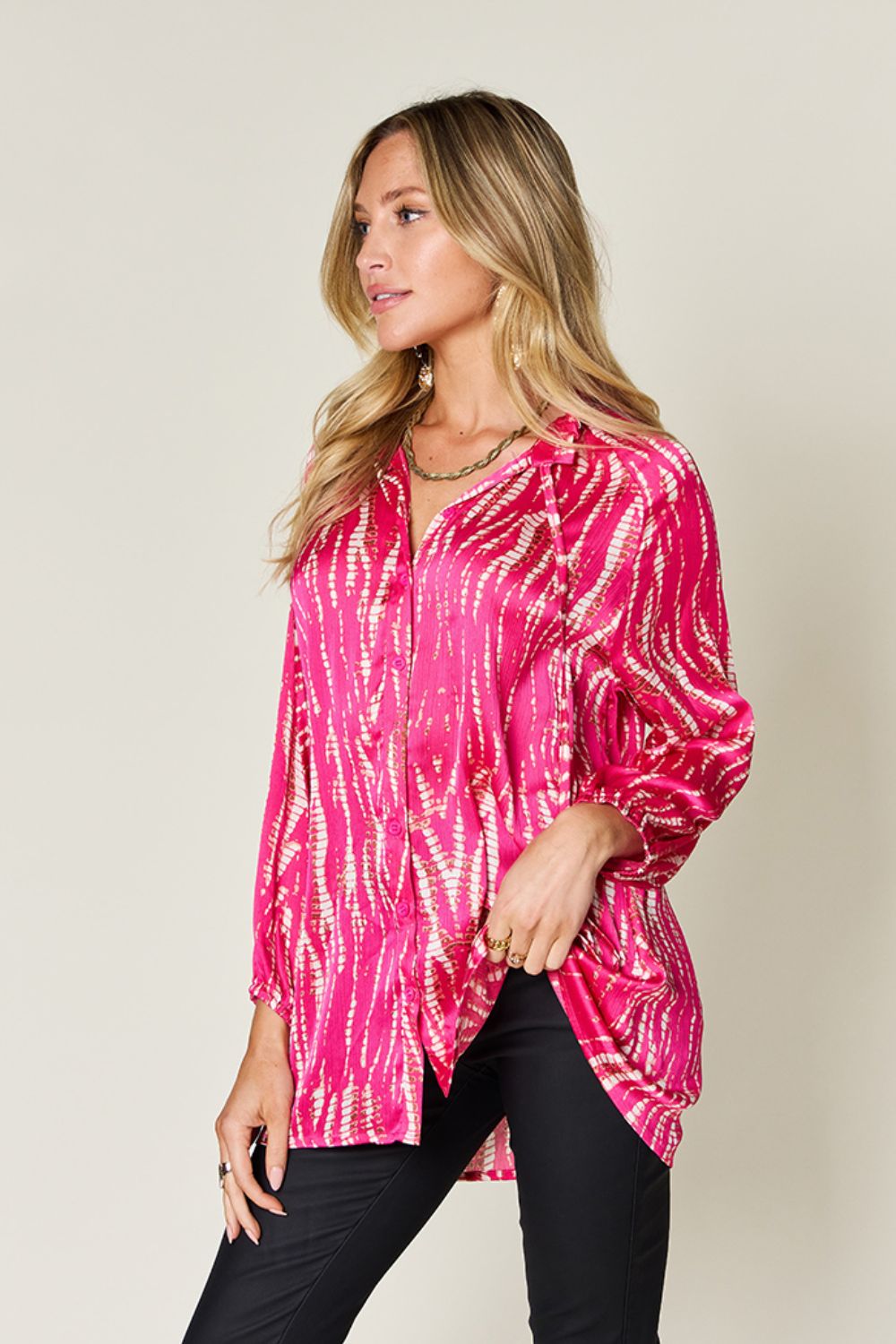 A woman wearing the Double Take Full Size Printed Button Up Long Sleeve Shirt in vibrant pink polyester and black pants stands against a plain background while touching her hair.