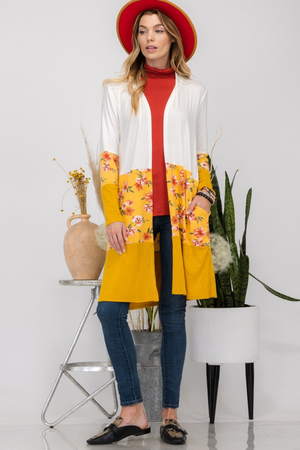 Wearing the Celeste Full Size Floral Color Block Open Front Cardigan with a white and yellow floral print, along with a red hat, a red top, and jeans, a woman stands beside a houseplant.