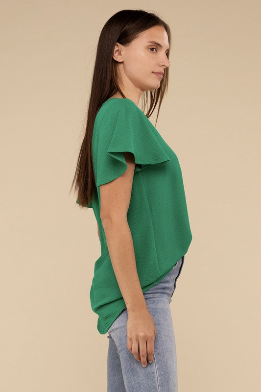 A woman with long straight hair models the Woven Airflow Flutter Sleeve Top in green, matched with light blue jeans, as she poses with her hands in her pockets against a plain beige background.