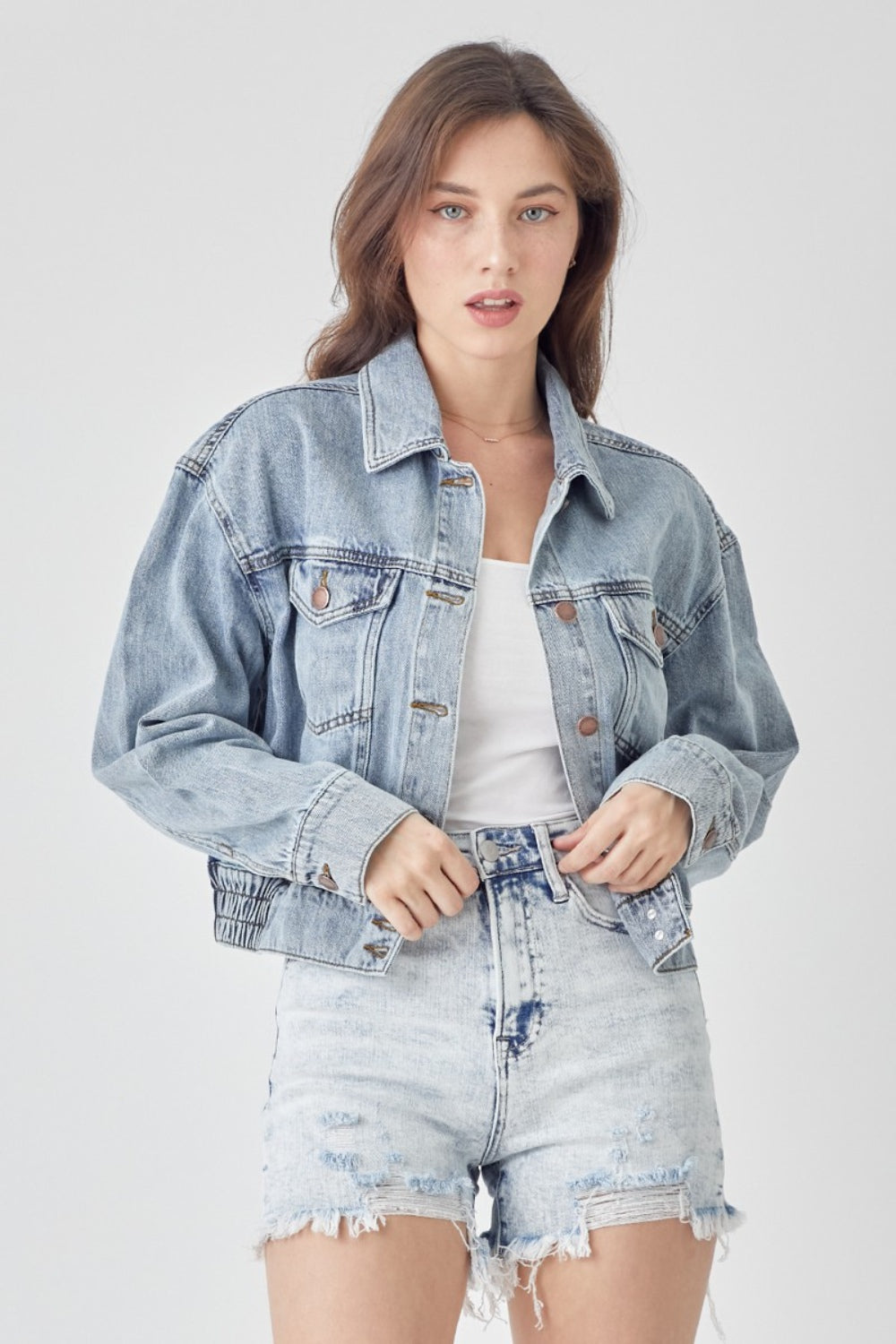 A woman wearing the RISEN Full Size Button Down Cropped Denim Jacket over a white top and distressed denim shorts stands against a plain background, showcasing versatile outerwear perfect for any casual occasion.