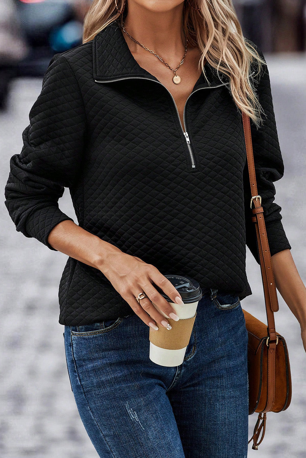 A person wearing a Black Solid Half Zipper Quilted Pullover Sweatshirt and blue jeans is walking down a cobblestone street while holding a gray clutch, embodying the essence of a casual wardrobe. The person's back is facing the camera.