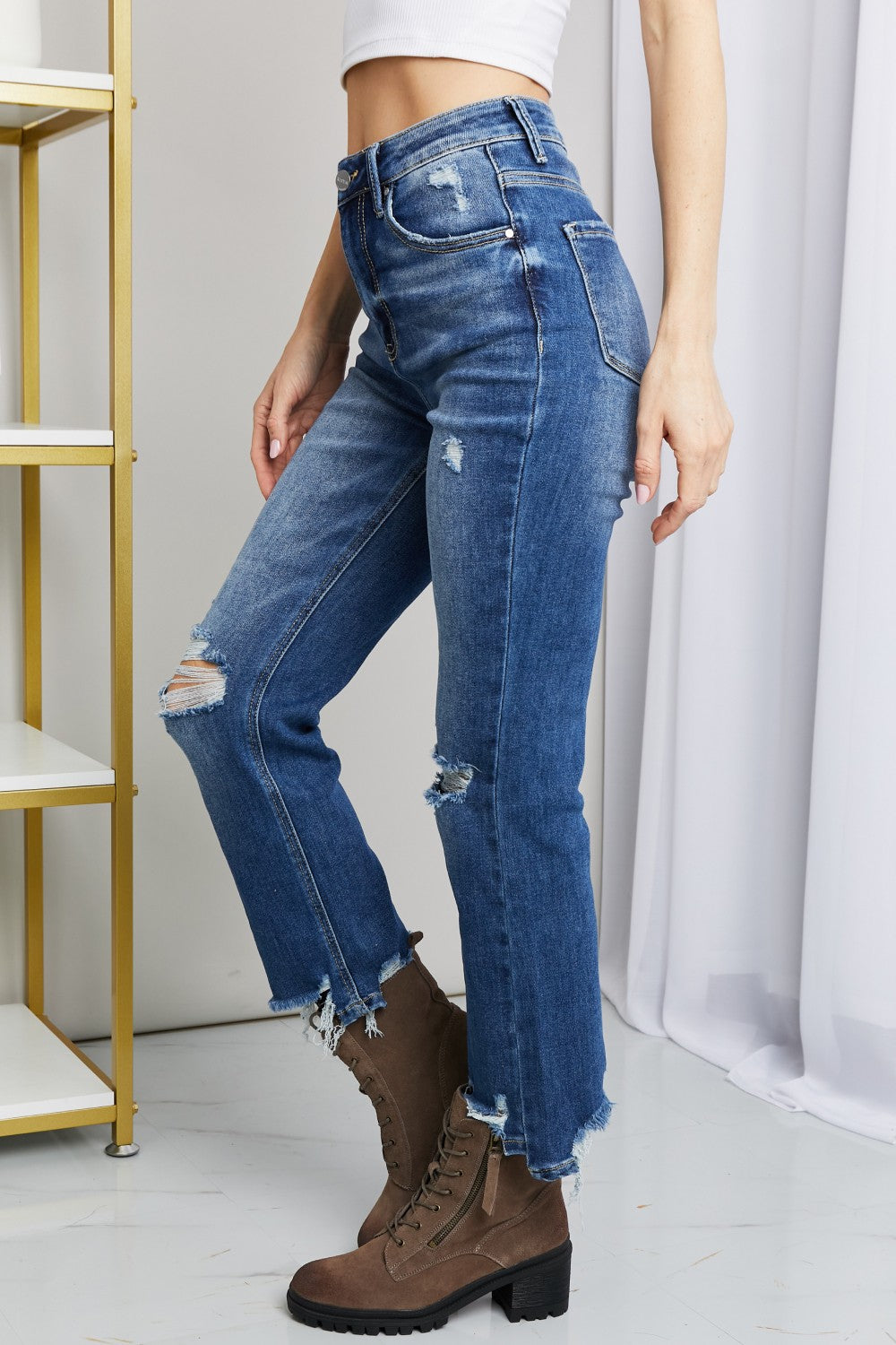 A person wearing RISEN Frayed Hem Distressed Cropped Jeans and brown ankle boots with a block heel stands in front of a white curtain next to a gold shelving unit.