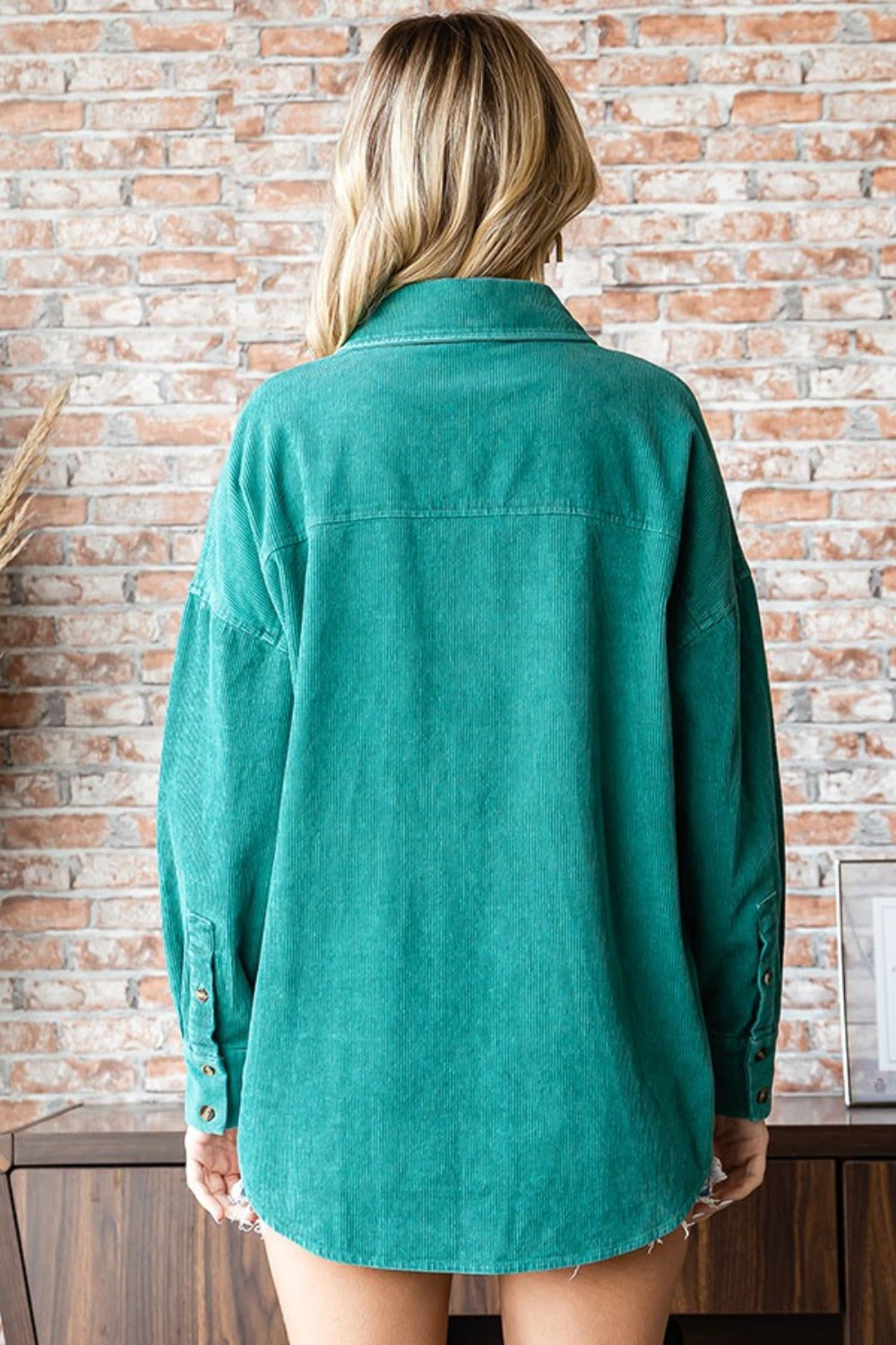 A woman wearing the First Love Washed Corduroy Button Down Long Sleeve Shirt in green, a white top, and denim shorts poses indoors. She stands in front of a brick wall with a leafy plant on the side.