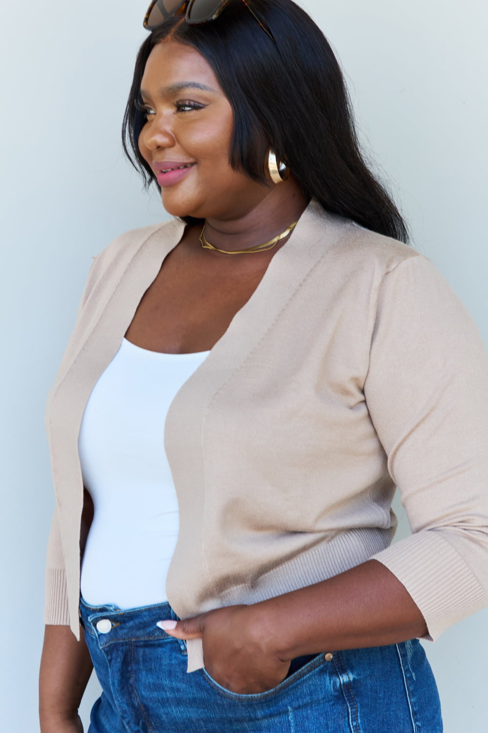 Woman with long dark hair wearing the versatile Doublju My Favorite Full Size 3/4 Sleeve Cropped Cardigan in Khaki over a white top and blue jeans, standing against a plain background.