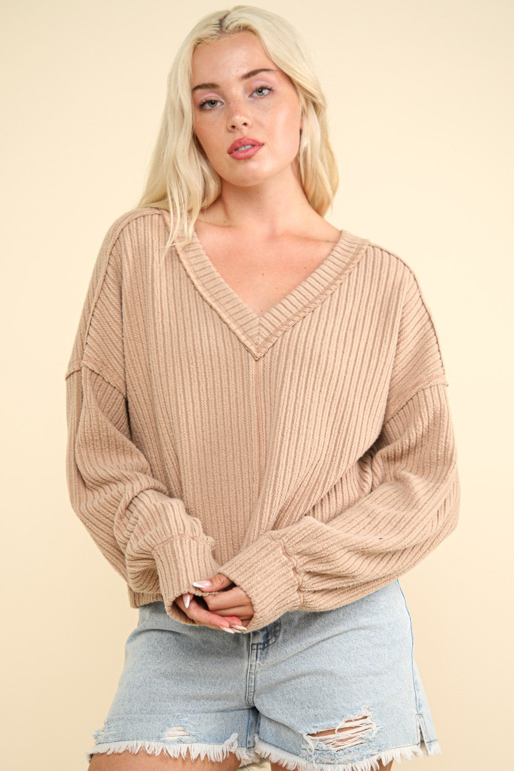 A woman with long blonde hair wears the VERY J Exposed Seam V-Neck Ribbed Knit Top, a casual and comfy piece featuring a ribbed knit texture and a drop shoulder design. She pairs it with light blue ripped denim shorts and stands against a cream-colored background.