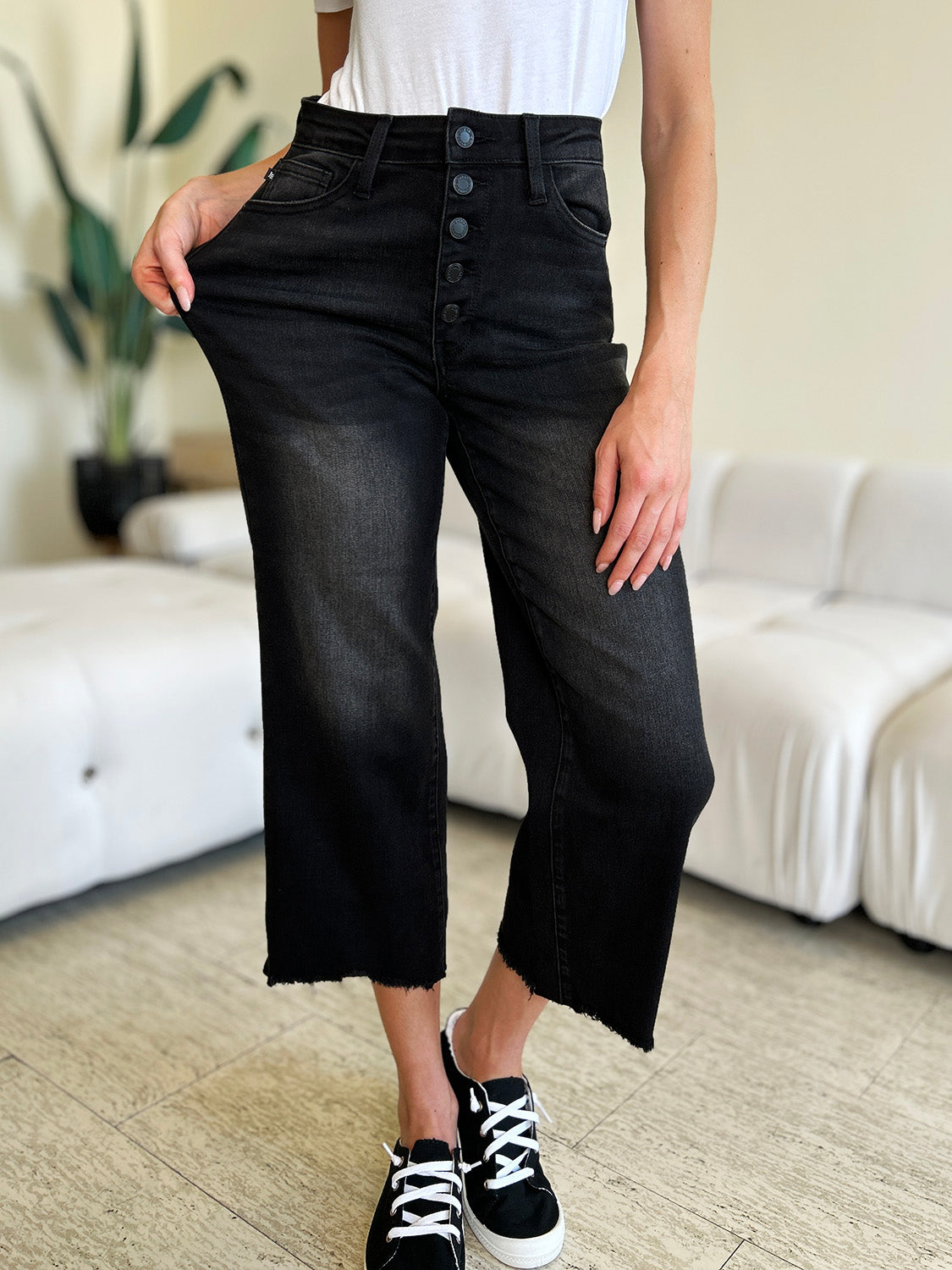 Person wearing Judy Blue Full Size High Waist Button Fly Jeans and black sneakers, standing indoors. Hands are in pockets, and the background includes a white couch and a plant. These high-waisted jeans make the outfit sleek and stylish.